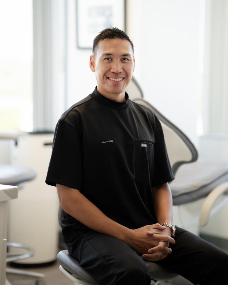 Dr. Aaron Ochoco, cosmetic dentist, smiling in black scrubs seated in a bright dental treatment room
